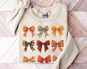 May include: A beige sweatshirt with a design of nine colorful bows in various patterns. The bows are in shades of orange, yellow, brown, and red.