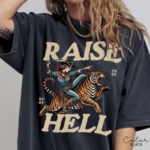 May include: Black graphic tee with a cowgirl riding a tiger, the words "RAISE HELL" and "STAY FREE" in a vintage style. The cowgirl wears a cowboy hat and holds a knife. The shirt is black.