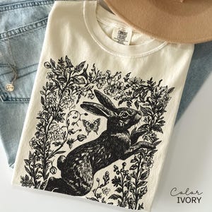 May include: Ivory-colored t-shirt featuring a black and white illustration of a rabbit surrounded by flowers and butterflies. The shirt has a crew neck and short sleeves. The text "Color IVORY" is in the bottom right corner.