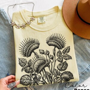 May include: A light yellow t-shirt with a black and white Venus flytrap graphic. The shirt is labeled "Comfort Colors" and the color is "Banana". A brown hat, a floral necklace, and blue jeans are also in the image.