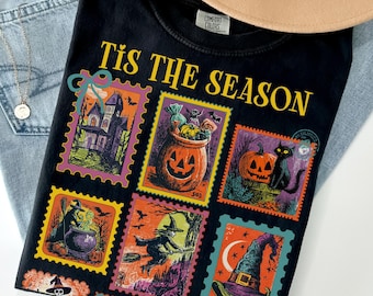 Może przedstawiać: Czarny t-shirt z motywem Halloween z wzorami znaczków pocztowych. Znaczki przedstawiają nawiedzony dom, dynie, czarnego kota, czarownicę i kapelusz czarownicy. Tekst "Tis the Season" jest wydrukowany u góry.