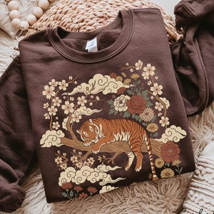 May include: Dark chocolate-colored sweatshirt featuring a tiger resting on a tree branch, surrounded by flowers and clouds. The design includes cream, brown, and red tones. The text "Color Dark Chocolate" is visible.