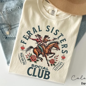 May include: Ivory t-shirt with a graphic of two cowgirls riding a horse. The graphic is surrounded by a circle with the text "Feral Sisters Social Club" and the words "Ride or Die" and "Sisters".