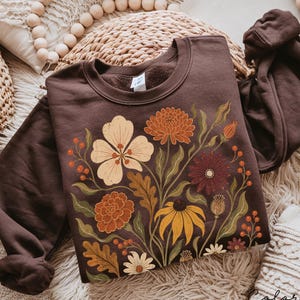 Puede incluir: Una sudadera de cuello redondo en color marr&oacute;n chocolate oscuro con un estampado gr&aacute;fico central de varias flores y follaje en tonos crema, naranja, amarillo y rojo intenso. El dise&ntilde;o incluye ilustraciones detalladas de crisantemos.