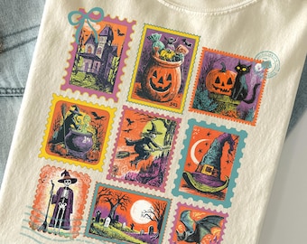 Puede incluir: Camiseta color marfil con diseños de sellos postales de Halloween. Los sellos representan casas encantadas, calabazas, brujas y murciélagos. La camiseta está doblada, con un sombrero y vaqueros en el fondo.
