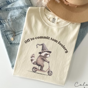 Könnte beinhalten: Ein cremefarbenes T-Shirt mit dem Text "Off to commit tom foolery" und einem Cartoon-Waschbären, der einen Hexenhut trägt und einen Roller fährt. Die Farbe des Shirts ist Elfenbein.