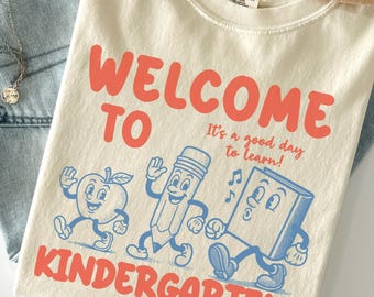 May include: Ivory-colored t-shirt with the words "WELCOME TO KINDERGARTEN" in coral lettering. The shirt features cartoon illustrations of an apple, pencil, and book. The text "It's a good day to learn!" is also printed on the shirt.