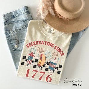 Puede incluir: Camiseta de color marfil con un gráfico de un perrito caliente, un fuego artificial y un vaso de soda. El gráfico es de estilo retro e incluye el texto "Celebrating Since 1776".