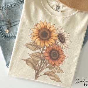 May include: A cream coloured t-shirt with a graphic of three sunflowers in shades of brown, orange, and yellow. The text "COMFORT COLORS" is visible on the tag.