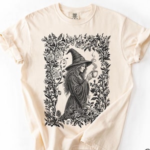 以下が含まれることがあります： アイボリー色のTシャツ。とんがり帽子と長いローブを身にまとい、湯気の立つカップを持つ人物の黒いインク画が描かれています。人物は様々な花や葉の密な枠で囲まれています。Color Ivoryというテキストが表示されています。
