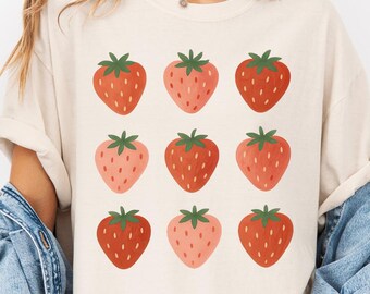 May include: Ivory-colored t-shirt featuring a grid of nine strawberries in varying shades of red and pink with green stems. The shirt is paired with a denim jacket. The text "Color Ivory" is displayed at the bottom left.
