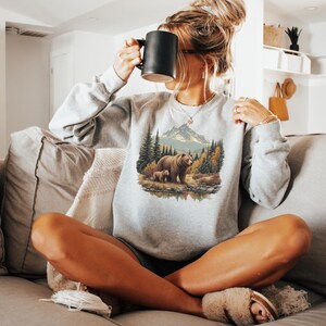 May include: Ash-colored sweatshirt featuring a nature scene with a bear and cub near a mountain and trees. The sweatshirt has long sleeves and a crew neck. The word "Color" and "Ash" are printed on the bottom right.