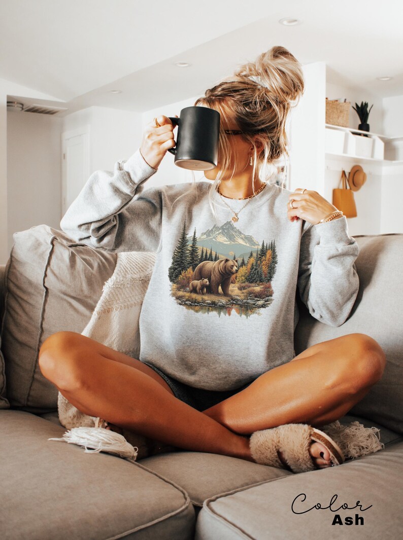 May include: Ash-colored sweatshirt featuring a nature scene with a bear and cub near a mountain and trees. The sweatshirt has long sleeves and a crew neck. The word "Color" and "Ash" are printed on the bottom right.