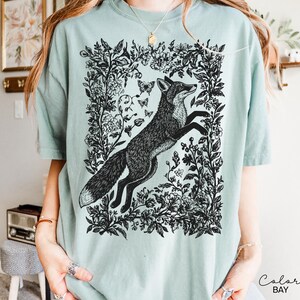 Pu&ograve; includere: T-shirt verde chiaro con un'illustrazione in bianco e nero di una volpe che salta all'interno di una cornice floreale. Il design include farfalle ed elementi botanici, creando un'estetica ispirata alla natura.