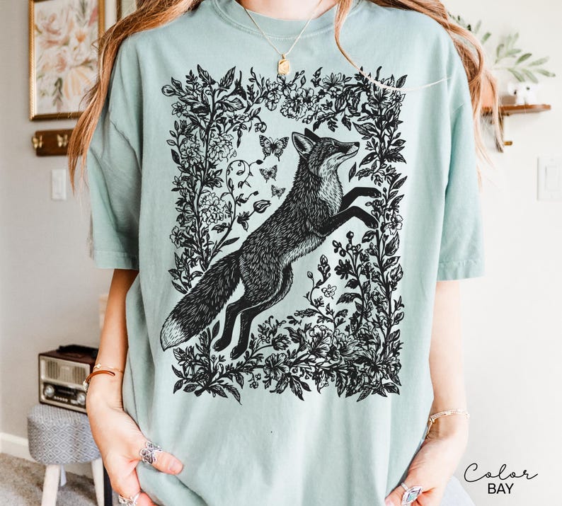 Pu&ograve; includere: T-shirt verde chiaro con un'illustrazione in bianco e nero di una volpe che salta all'interno di una cornice floreale. Il design include farfalle ed elementi botanici, creando un'estetica ispirata alla natura.
