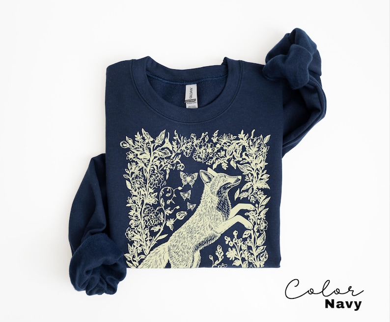 Pu&ograve; includere: Felpa blu navy con un'illustrazione di volpe color crema circondata da elementi floreali e farfalle. Il testo "Color Navy" &egrave; scritto in corsivo in basso a destra. La felpa &egrave; uno stile girocollo.