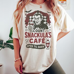 May include: Ivory-colored t-shirt featuring a graphic of Count Snackula's Cafe. The design includes a cartoon vampire holding a milkshake, bats, and the text "OPEN TIL DAWN". The shirt is a comfortable, casual style.