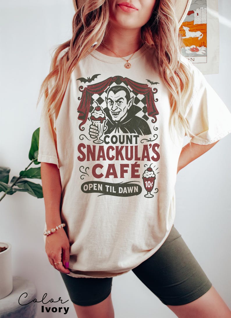May include: Ivory-colored t-shirt featuring a graphic of Count Snackula's Cafe. The design includes a cartoon vampire holding a milkshake, bats, and the text "OPEN TIL DAWN". The shirt is a comfortable, casual style.