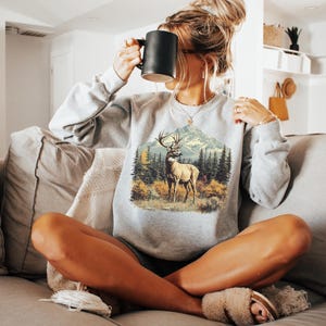 May include: Ash-colored sweatshirt featuring a detailed graphic of a deer in a forest scene with mountains in the background. The sweatshirt has long sleeves and a crew neck. The word "Color" and "Ash" are printed at the bottom of the image.