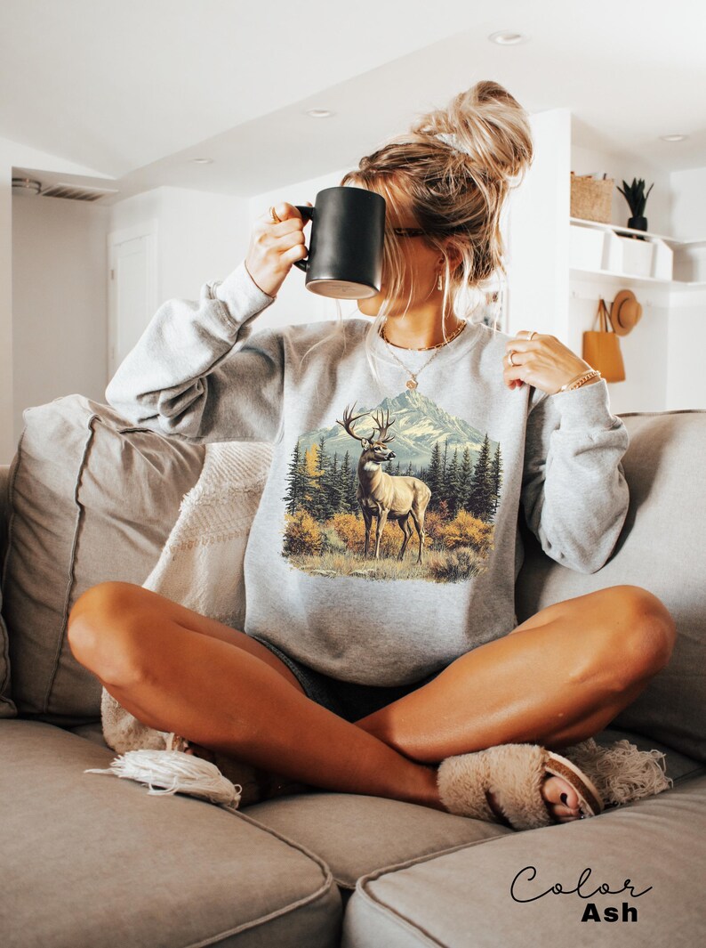 May include: Ash-colored sweatshirt featuring a detailed graphic of a deer in a forest scene with mountains in the background. The sweatshirt has long sleeves and a crew neck. The word "Color" and "Ash" are printed at the bottom of the image.