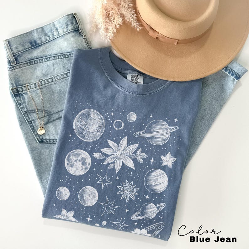 Puede incluir: Una camiseta azul jean con un estampado blanco de planetas, estrellas y flores. El texto "Comfy Colors" es visible en la etiqueta.