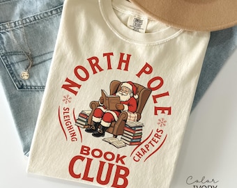 May include: Ivory-colored t-shirt with a red graphic that reads "North Pole Book Club." The graphic features Santa Claus reading in a chair, surrounded by books. The shirt is folded on top of blue jeans and a tan hat.