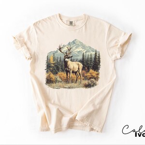 May include: Ivory-colored t-shirt featuring a detailed graphic of a deer in a forest scene with mountains in the background. The deer has large antlers and is standing in a grassy area. The shirt has a classic crew neck and short sleeves.