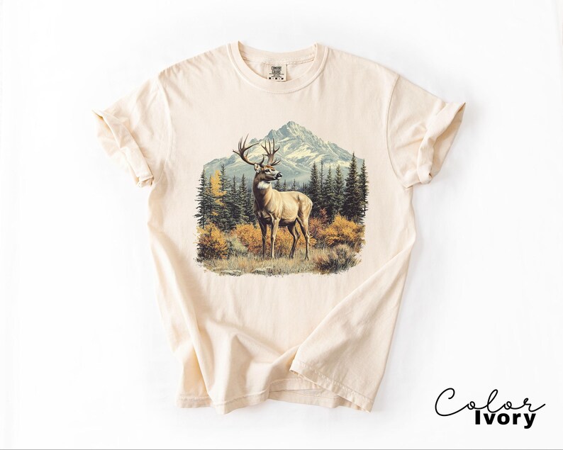 May include: Ivory-colored t-shirt featuring a detailed graphic of a deer in a forest scene with mountains in the background. The deer has large antlers and is standing in a grassy area. The shirt has a classic crew neck and short sleeves.