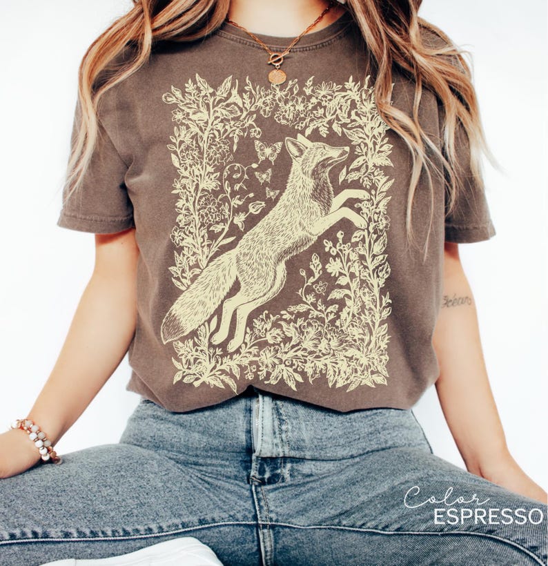 Pu&ograve; includere: Una maglietta marrone con un'illustrazione color crema di una volpe che salta all'interno di una cornice floreale. Il colore della maglietta &egrave; etichettato "Color Espresso" in basso a destra. Il design include farfalle ed elementi botanici.
