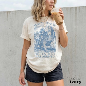 May include: Ivory-colored t-shirt with a blue graphic of a knight riding a tiger, with the text "HEAR YE! HEAR YE!" above and "THE FUN AUNT HAS ARRIVED" below. The shirt is worn by a person.