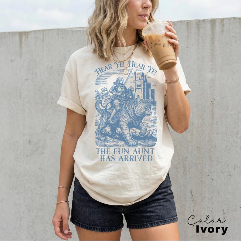 May include: Ivory-colored t-shirt with a blue graphic of a knight riding a tiger, with the text "HEAR YE! HEAR YE!" above and "THE FUN AUNT HAS ARRIVED" below. The shirt is worn by a person.