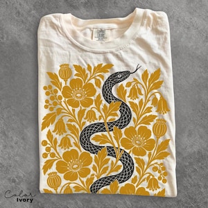 Può includere: Una t-shirt color avorio con una stampa grafica di un serpente nero che si snoda attraverso un denso arrangiamento di fiori, foglie e baccelli color giallo senape. La t-shirt ha un girocollo e un'etichetta Comfort Colors sul colletto.