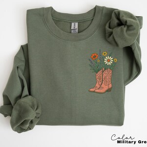Puede incluir: Sudadera verde militar con dise&ntilde;o floral. El gr&aacute;fico presenta un par de botas de vaquero de color coral llenas de flores silvestres. El texto "Color Military Green" est&aacute; en la parte inferior derecha.