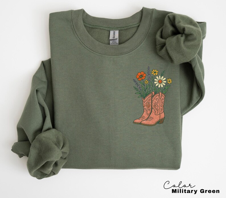 Puede incluir: Sudadera verde militar con dise&ntilde;o floral. El gr&aacute;fico presenta un par de botas de vaquero de color coral llenas de flores silvestres. El texto "Color Military Green" est&aacute; en la parte inferior derecha.