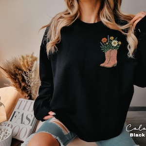 Puede incluir: Sudadera negra con un gr&aacute;fico de botas de vaquero rosas llenas de flores coloridas. Las botas tienen detalles intrincados. Las palabras "Color" y "Black" est&aacute;n escritas en cursiva en la esquina inferior derecha.