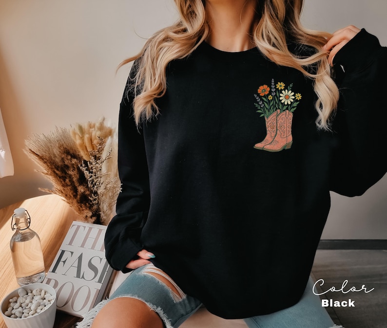 Puede incluir: Sudadera negra con un gr&aacute;fico de botas de vaquero rosas llenas de flores coloridas. Las botas tienen detalles intrincados. Las palabras "Color" y "Black" est&aacute;n escritas en cursiva en la esquina inferior derecha.