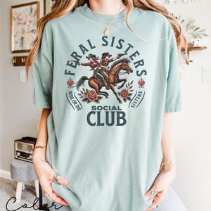 May include: A light blue t-shirt with a graphic of two women riding a horse. The graphic includes the text "Feral Sisters Social Club" and "Ride or Die".
