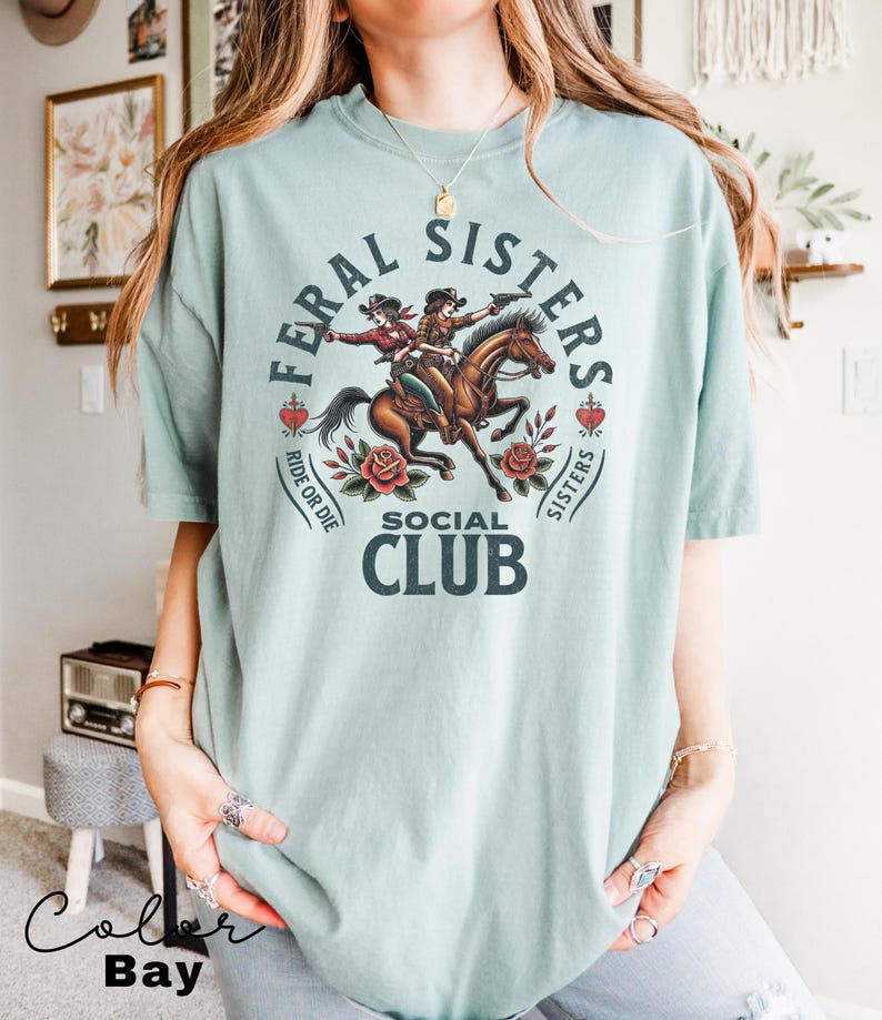 May include: A light blue t-shirt with a graphic of two women riding a horse. The graphic includes the text "Feral Sisters Social Club" and "Ride or Die".