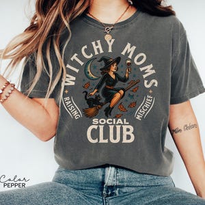 Puede incluir: Camiseta color pimienta con un gr&aacute;fico que dice "Witchy Moms Social Club". El dise&ntilde;o presenta una bruja montada en una escoba, un gato negro, una luna creciente y hojas de oto&ntilde;o. La camiseta es de corte holgado.