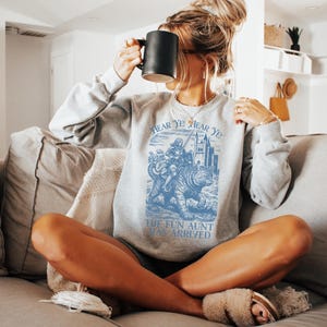May include: Ash-colored sweatshirt with a blue graphic that reads "Hear Ye! Hear Ye! The Fun Aunt Has Arrived." The graphic depicts a person riding a tiger. The sweatshirt is long-sleeved and has a crew neck.