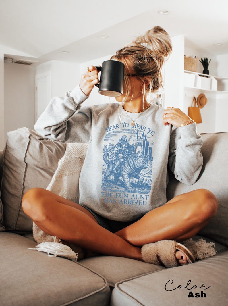May include: Ash-colored sweatshirt with a blue graphic that reads "Hear Ye! Hear Ye! The Fun Aunt Has Arrived." The graphic depicts a person riding a tiger. The sweatshirt is long-sleeved and has a crew neck.