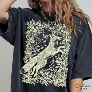 Pu&ograve; includere: T-shirt nero con un'illustrazione color crema di una volpe che salta all'interno di un disegno floreale e di farfalle. Il testo "Color BLACK" si trova nell'angolo in basso a destra. La maglietta ha maniche corte e una vestibilit&agrave; ampia.