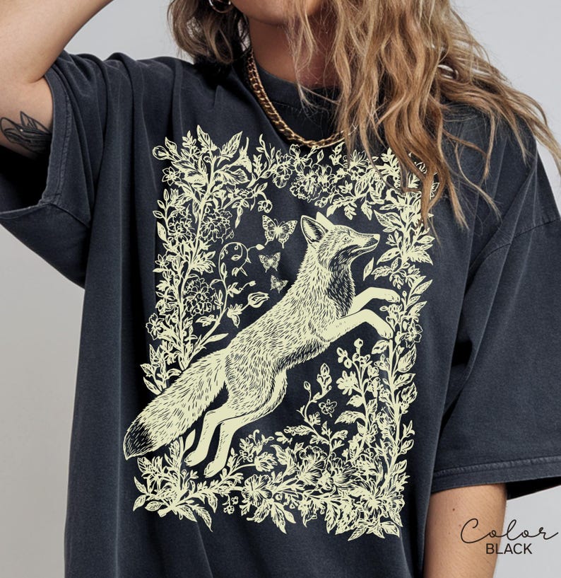 Pu&ograve; includere: T-shirt nero con un'illustrazione color crema di una volpe che salta all'interno di un disegno floreale e di farfalle. Il testo "Color BLACK" si trova nell'angolo in basso a destra. La maglietta ha maniche corte e una vestibilit&agrave; ampia.