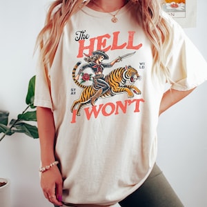 May include: A cream colored t-shirt with a graphic of a woman riding a tiger. The text on the shirt reads "The Hell I Won't".