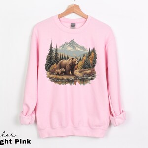 May include: Light pink sweatshirt featuring a detailed illustration of a mother bear and cub in a forest setting. The design includes trees, a mountain range, and a body of water. The text "Color Light Pink" is at the bottom.