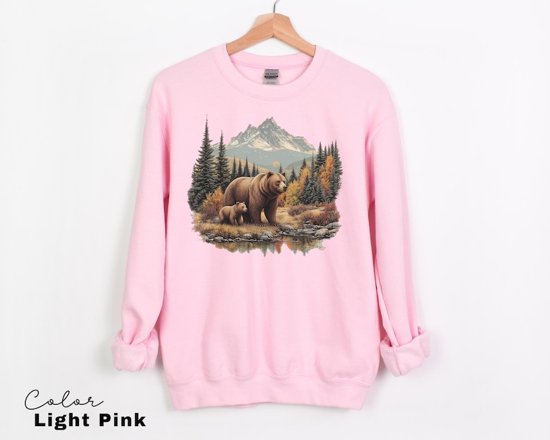 May include: Light pink sweatshirt featuring a detailed illustration of a mother bear and cub in a forest setting. The design includes trees, a mountain range, and a body of water. The text "Color Light Pink" is at the bottom.