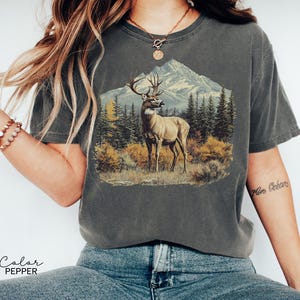 May include: Dark gray t-shirt featuring a detailed illustration of a deer in a forest setting with a mountain backdrop. The graphic includes trees and foliage in shades of green, yellow, and brown. The text "Color Pepper" is at the bottom.