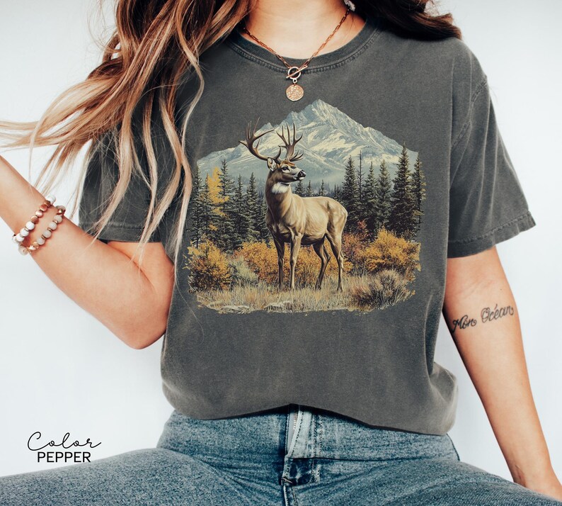 May include: Dark gray t-shirt featuring a detailed illustration of a deer in a forest setting with a mountain backdrop. The graphic includes trees and foliage in shades of green, yellow, and brown. The text "Color Pepper" is at the bottom.