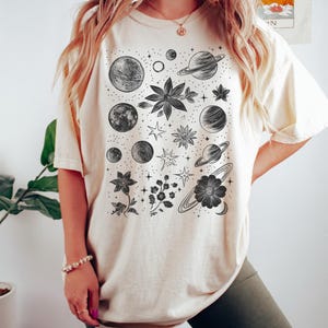 Puede incluir: Una camiseta de color crema con un estampado gr&aacute;fico en blanco y negro de planetas, estrellas y flores. El texto "Color Ivory" est&aacute; en la parte inferior de la imagen.