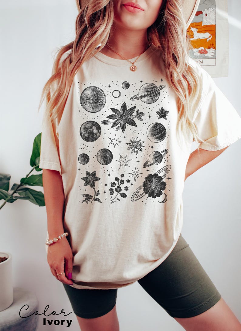 Puede incluir: Una camiseta de color crema con un estampado gr&aacute;fico en blanco y negro de planetas, estrellas y flores. El texto "Color Ivory" est&aacute; en la parte inferior de la imagen.